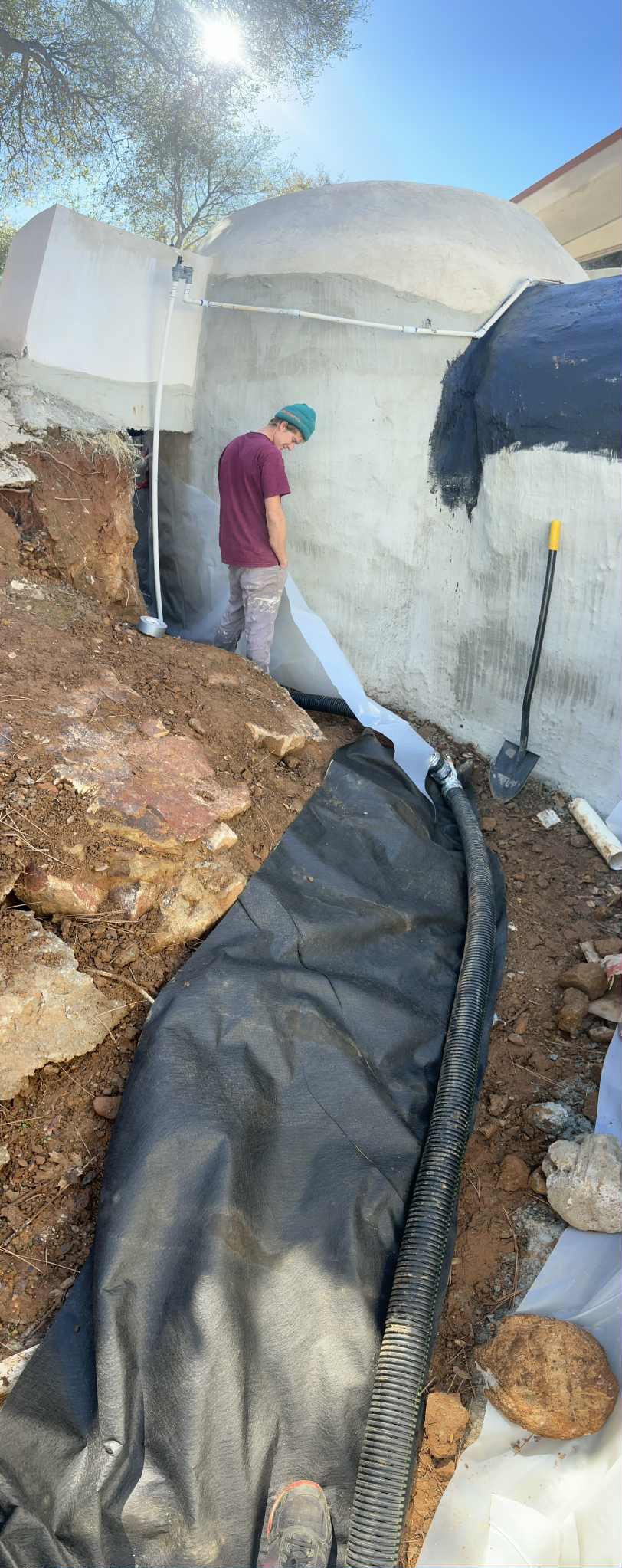 A builder standing beside a white ferro-cement dome wall, with corrugated drain pipe and filter fabric laid along the trench