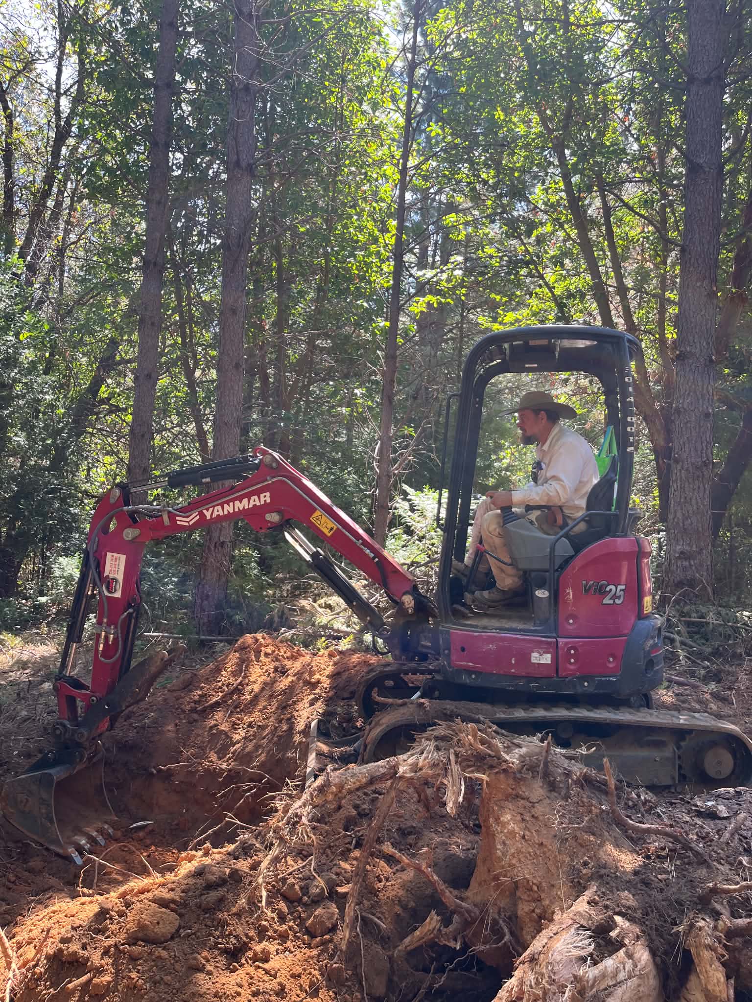 A guild member in a wide-brimmed hat operating a red mini excavator, digging into red soil among pines