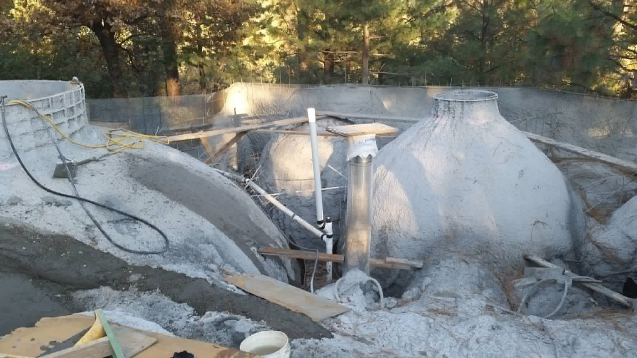 Several onion-shaped ferro-cement domes freshly sprayed with gray cement, with pipes and boards between them in a pine forest