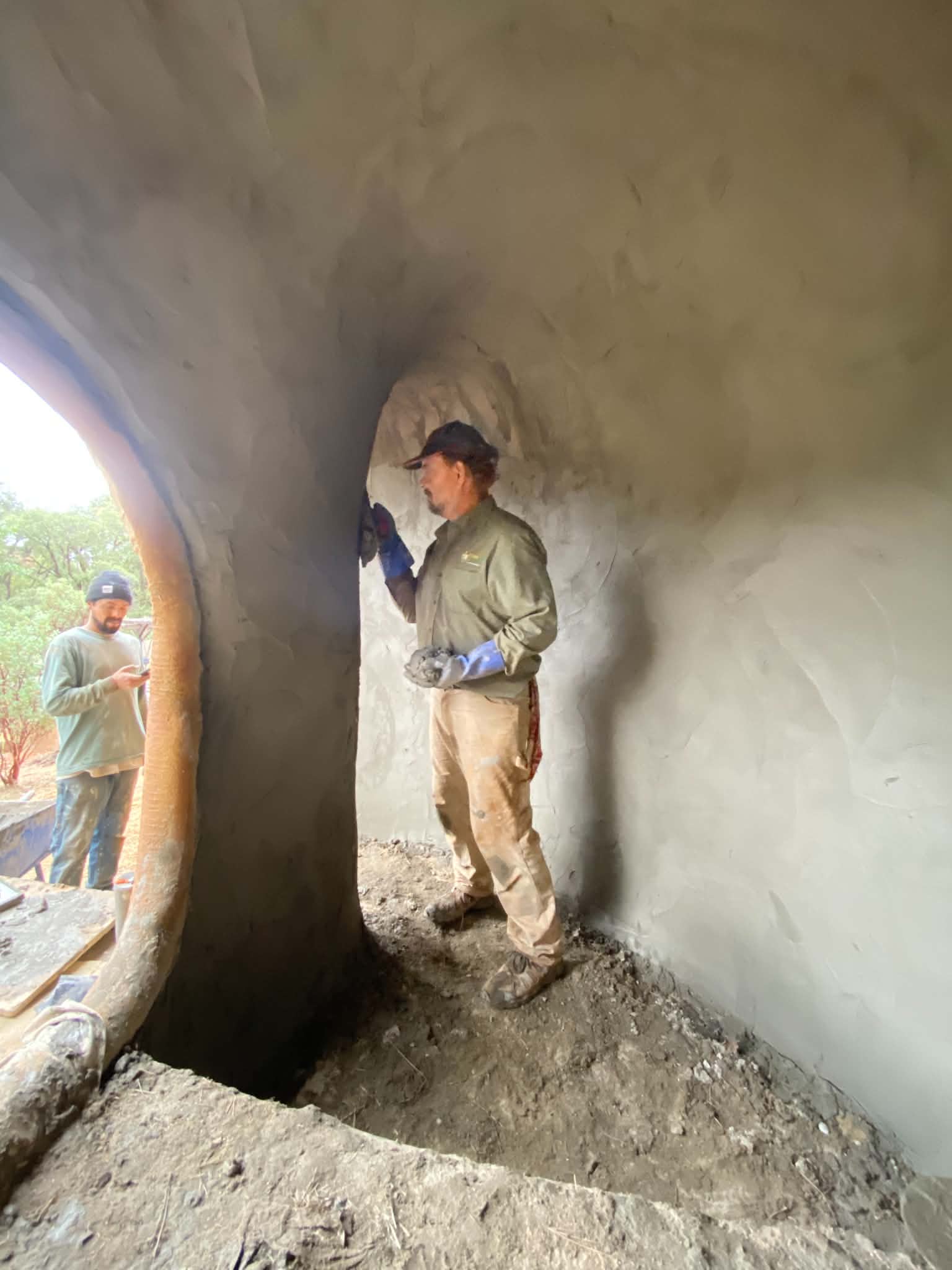 A craftsman in gloves hand-packing cement onto the curved interior wall of a ferro-cement room beside an arched opening
