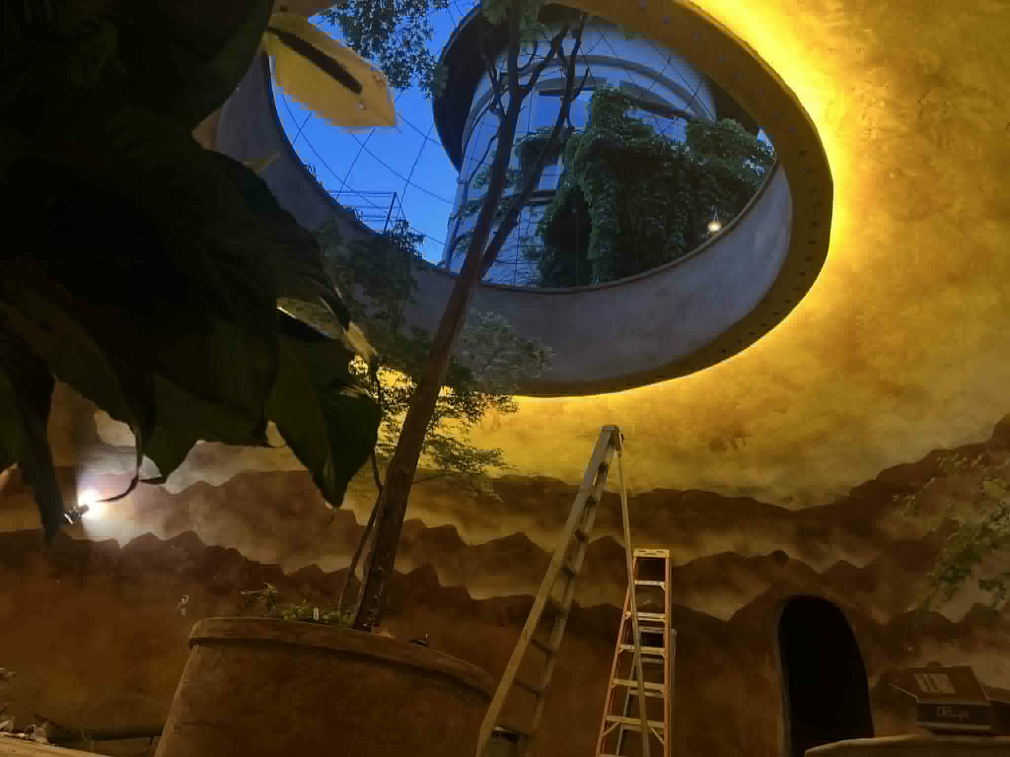 Looking up through a glowing oculus skylight in a gold-washed ferro-cement ceiling above a painted mountain mural