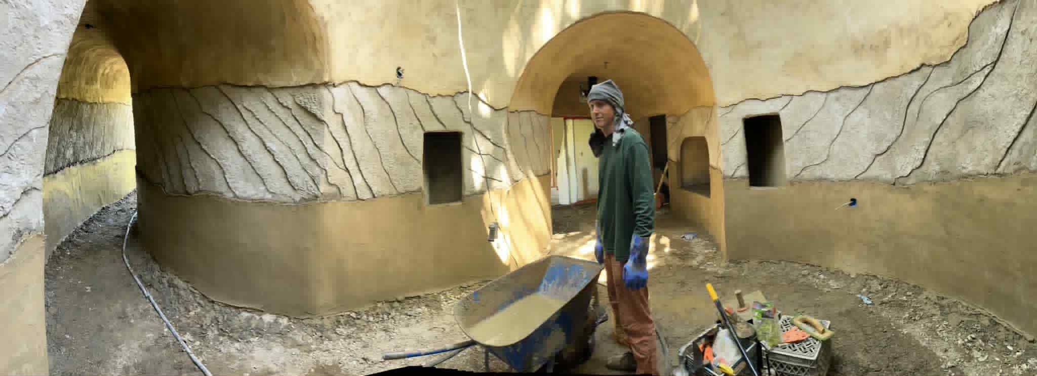 Panorama of a round ferro-cement room with a carved stone-texture band, arched passages, and a builder beside a wheelbarrow