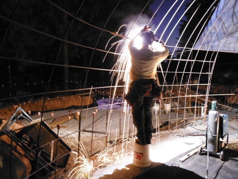 A builder standing on a bucket welding steel arch hoops at night, sparks showering down