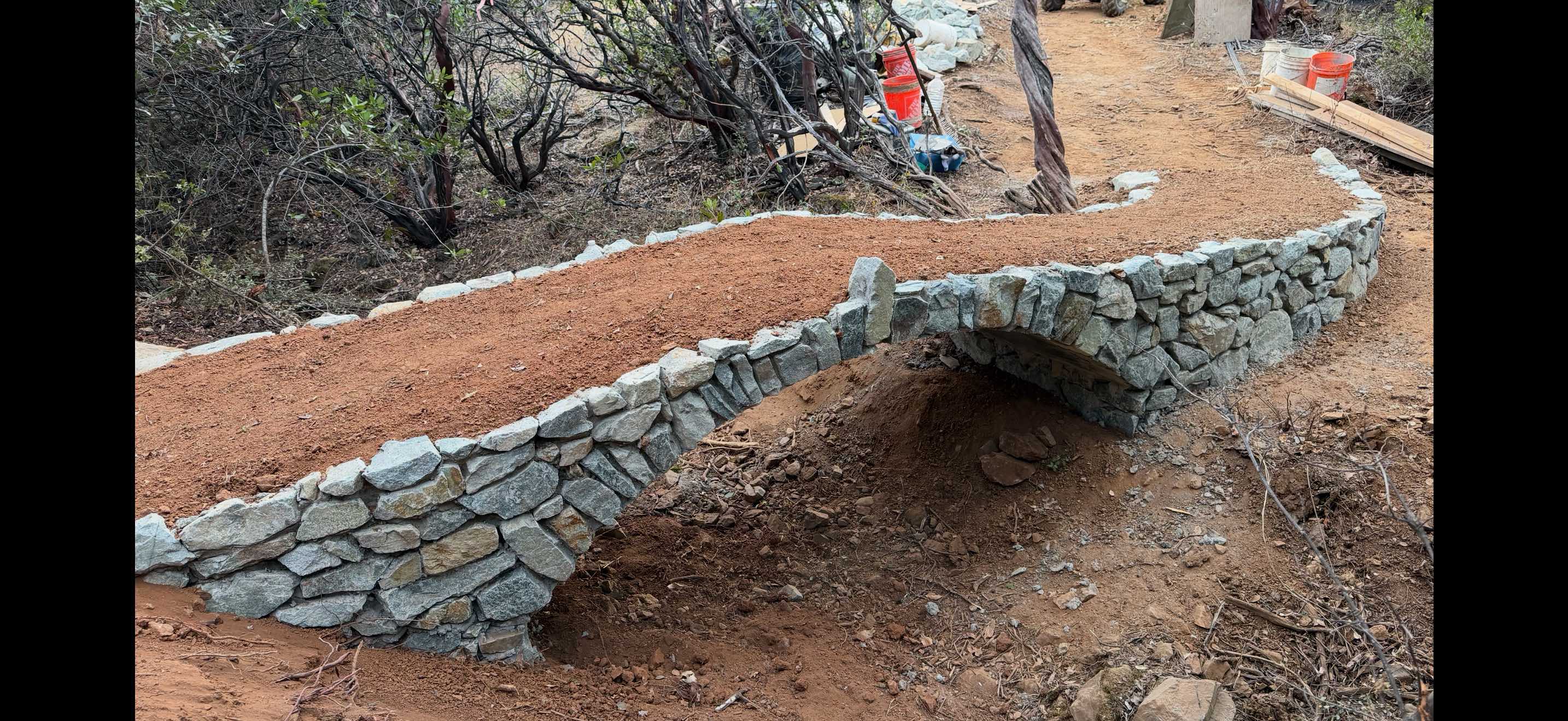 A finished stone arch bridge with a red-earth deck and curving dry-stone walls on a foothill trail