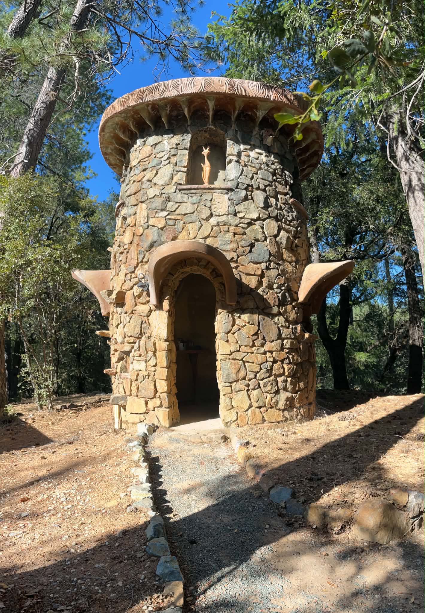 A round fieldstone tower with a sculpted cement mushroom-cap roof and arched doorway, set among pines