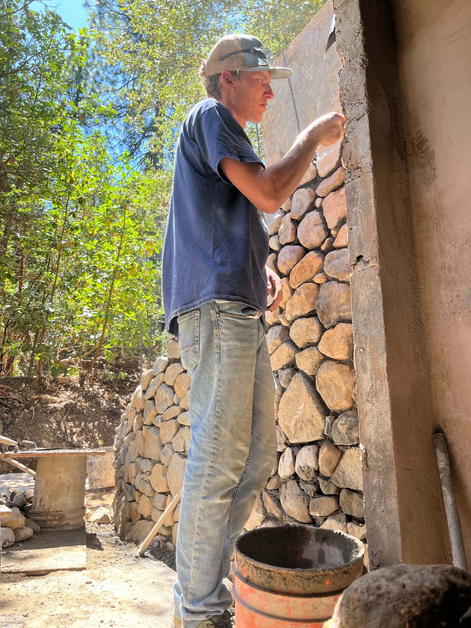 A guild volunteer setting river stones into a mortared wall beside a ferro-cement form